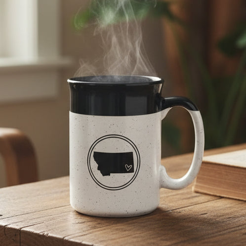 White mug with black interior and handle, featuring a black outline of a state with a heart, on a wooden surface with a grassy field background.