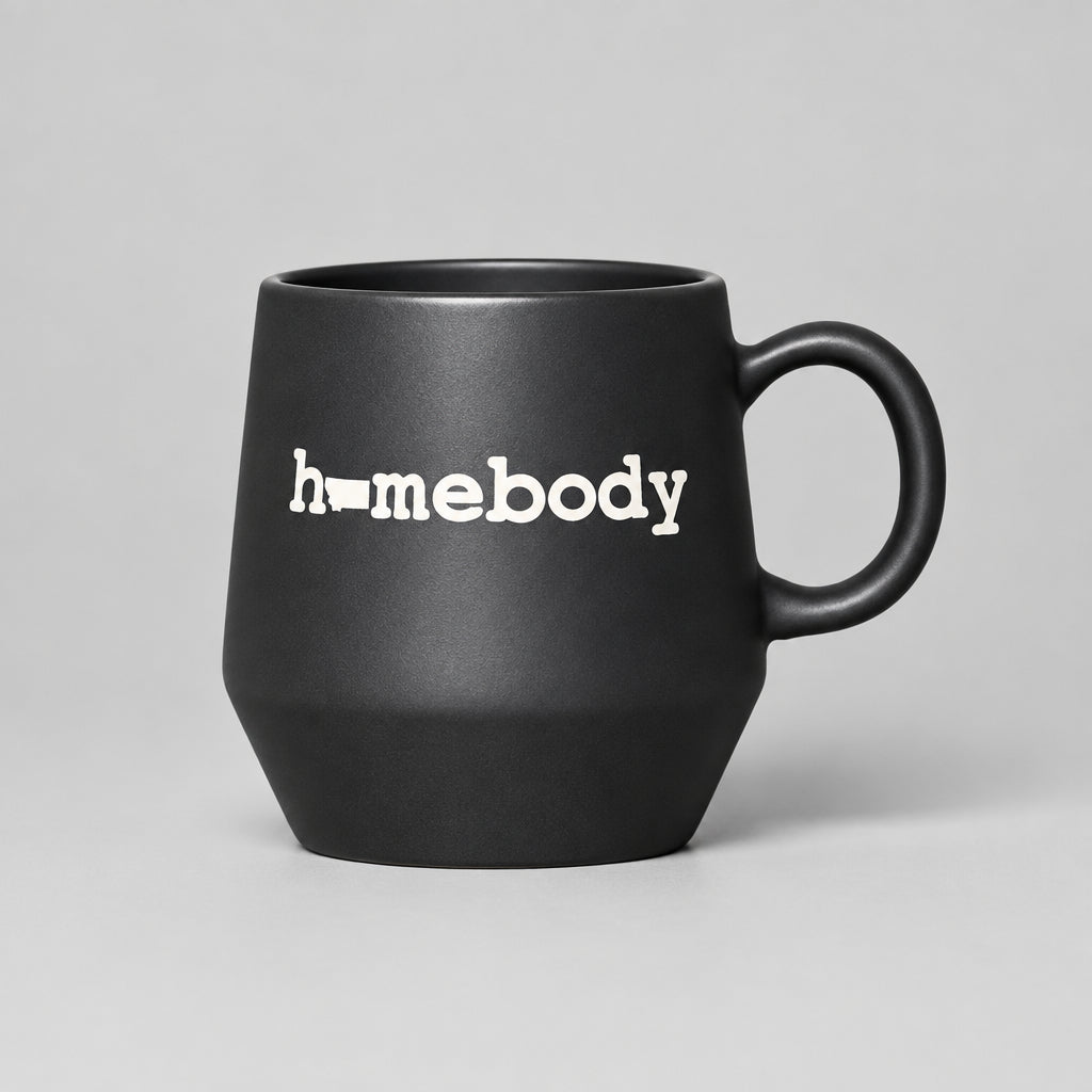 A matte black ceramic mug with the word 'homebody' printed in white lowercase letters, resting on a weathered wooden log with a blurred field of dry grass in the background.