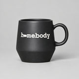 A matte black ceramic mug with the word 'homebody' printed in white lowercase letters, resting on a weathered wooden log with a blurred field of dry grass in the background.