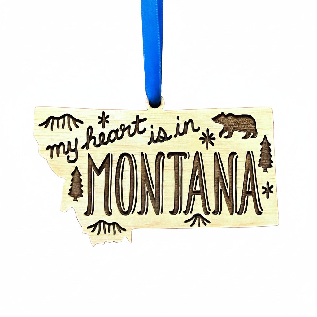 A wooden ornament shaped like the state of Montana, with the phrase "my heart is in MONTANA" engraved across the front, along with a bear, trees, and stars. The ornament hangs from a bright blue ribbon against a plain white background.