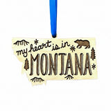 A wooden ornament shaped like the state of Montana, with the phrase "my heart is in MONTANA" engraved across the front, along with a bear, trees, and stars. The ornament hangs from a bright blue ribbon against a plain white background.