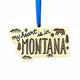 A wooden ornament shaped like the state of Montana, with the phrase "my heart is in MONTANA" engraved across the front, along with a bear, trees, and stars. The ornament hangs from a bright blue ribbon against a plain white background.