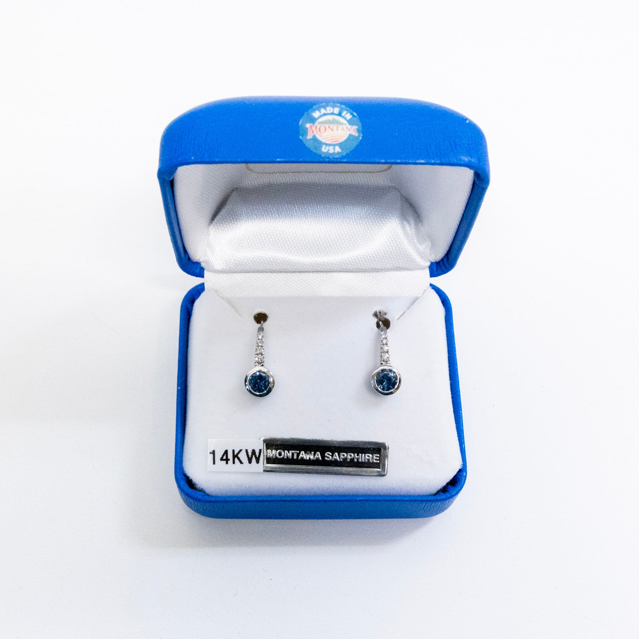 A pair of 14KW Montana Sapphire drop earrings displayed in an open blue jewelry box with a white interior. The earrings feature round blue sapphires and silver-toned metal settings.
