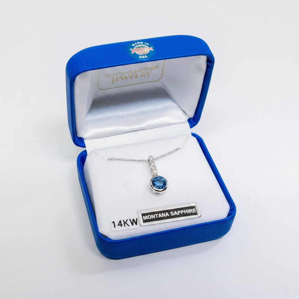 A silver pendant necklace with a round-cut blue sapphire gemstone, presented in an open blue jewelry box with a white interior. The pendant is labeled with a silver tag reading '14KW MONTANA SAPPHIRE'.