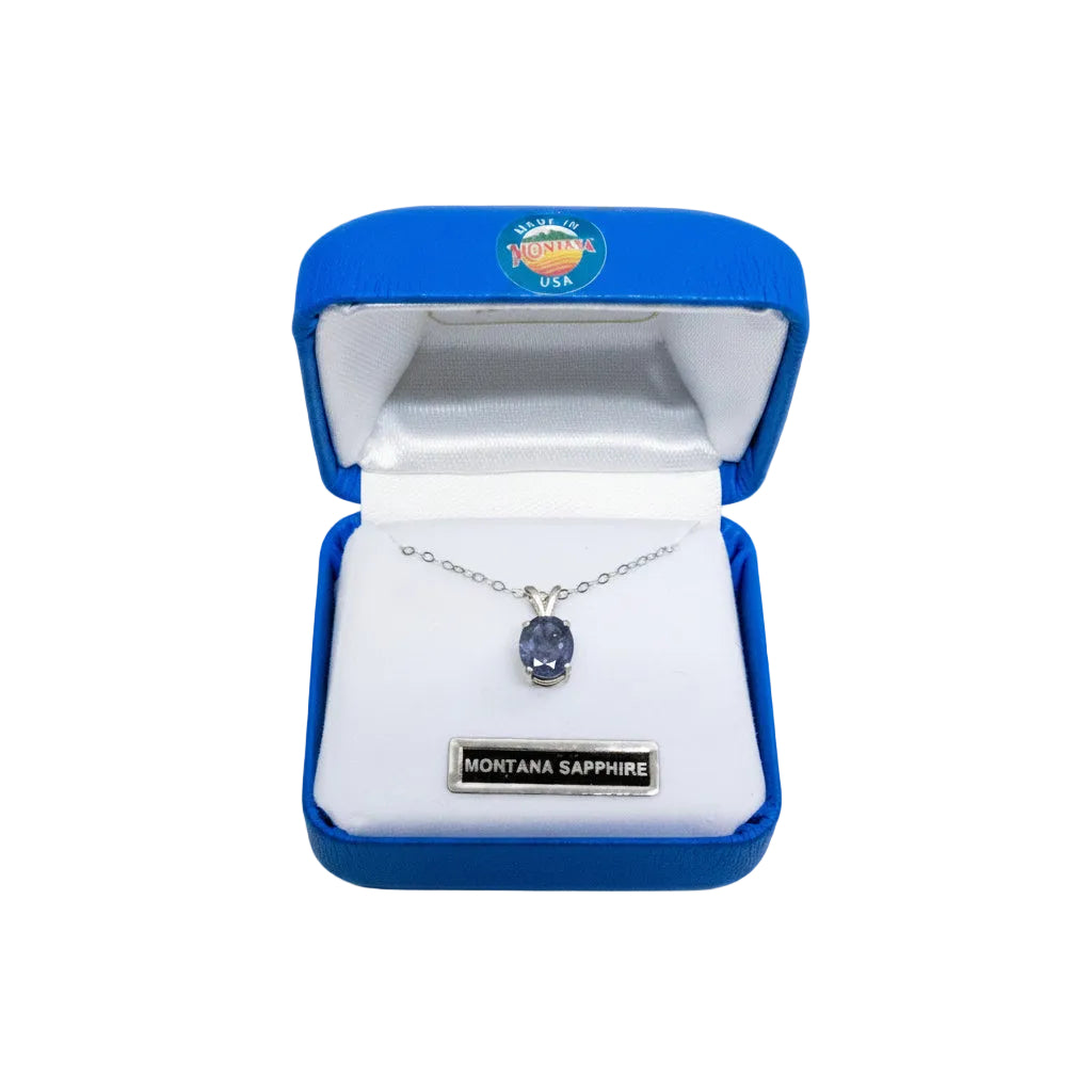 A silver necklace with a round Montana Sapphire pendant displayed inside an open blue jewelry box with a white interior. The box has a Montana Sapphire USA logo on the inside lid and a silver brand plaque at the bottom.