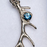 A close-up of a silver branch-shaped pendant featuring a round, faceted blue gemstone in a prong setting, shown against a plain white background.