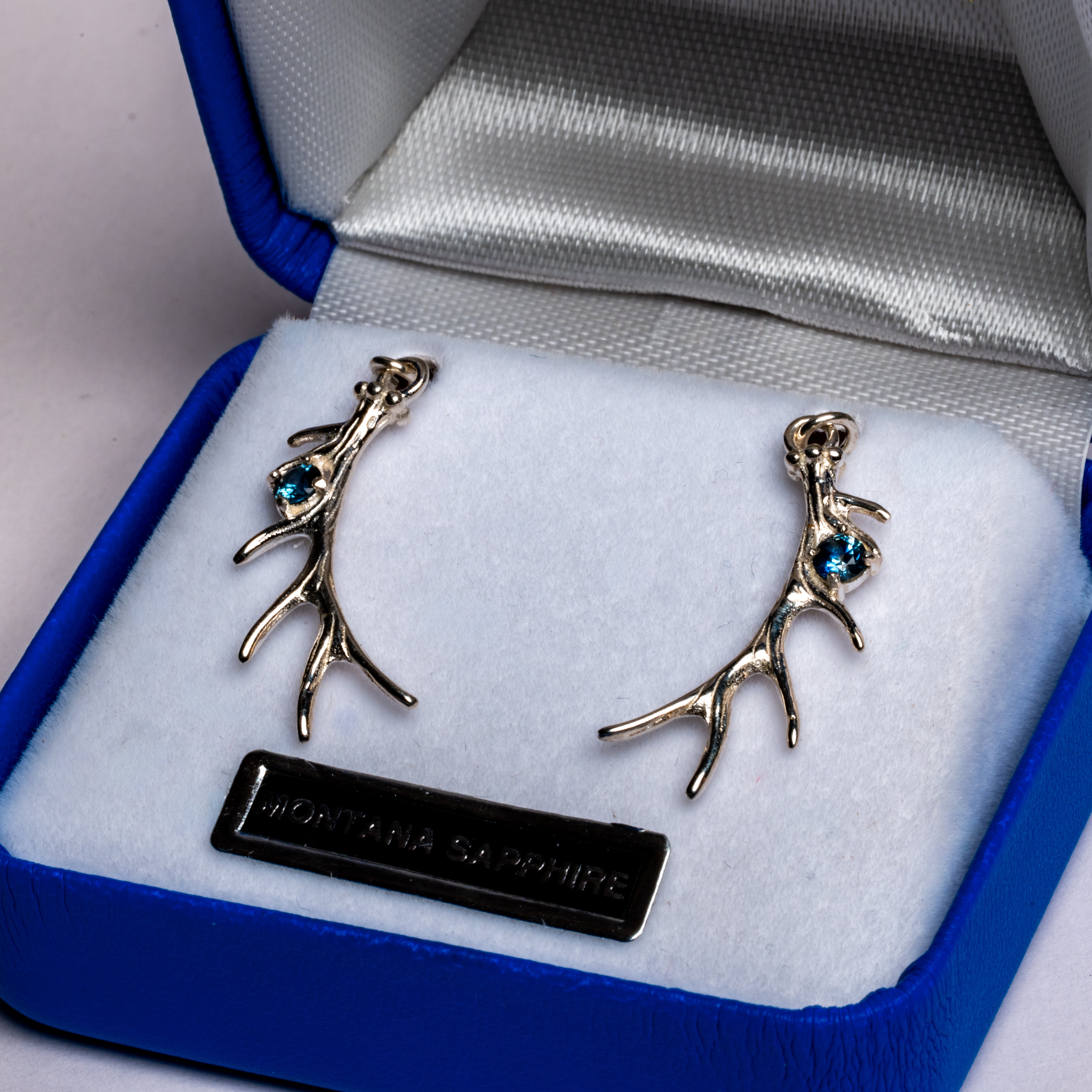 A pair of silver-toned antler stud earrings with small blue gemstone accents, displayed in an open blue jewelry box with a white cushion and a black brand plaque reading 'Montana Sapphire'.
