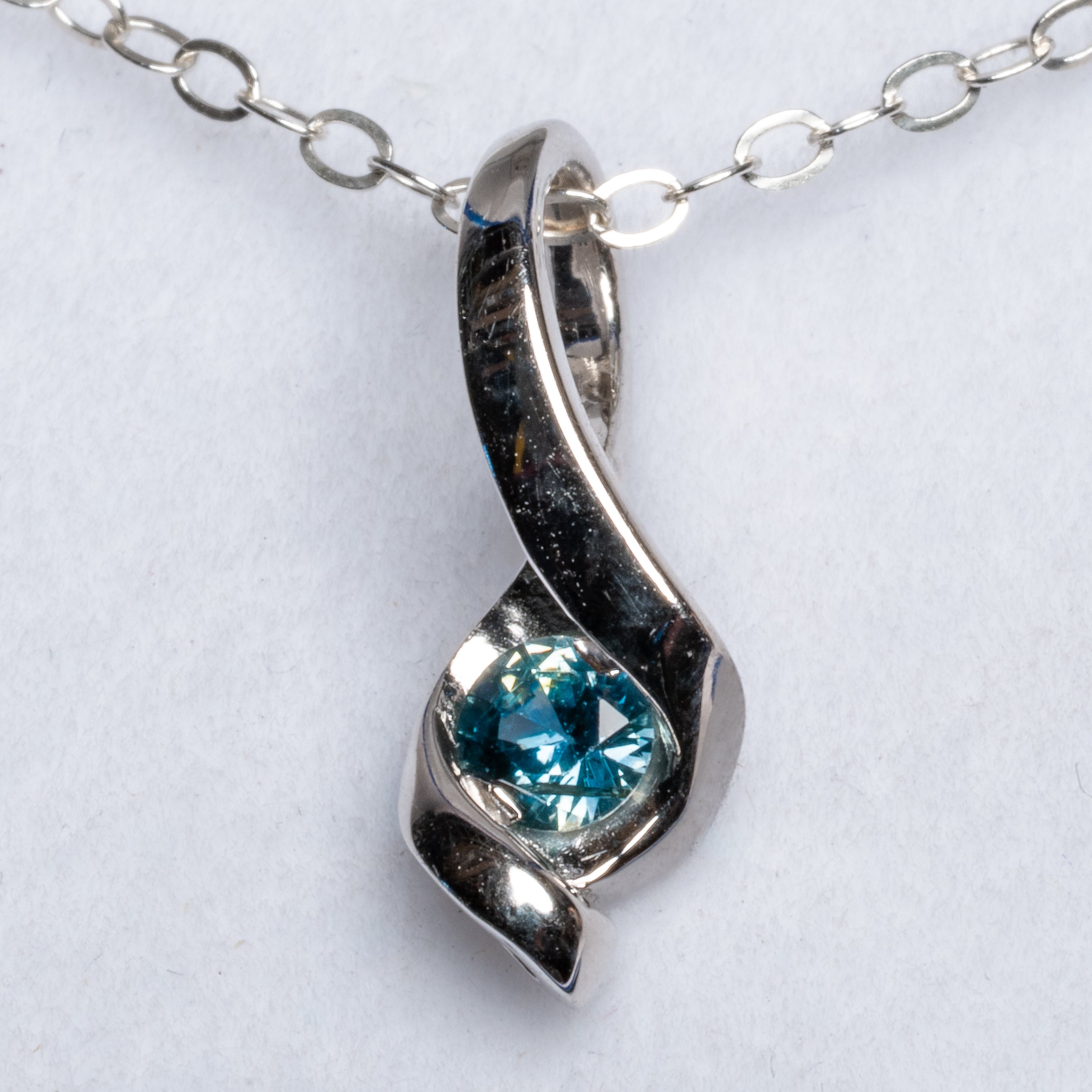 A silver pendant necklace featuring a twisted ribbon design with a round faceted blue gemstone, shown on a white textured background.