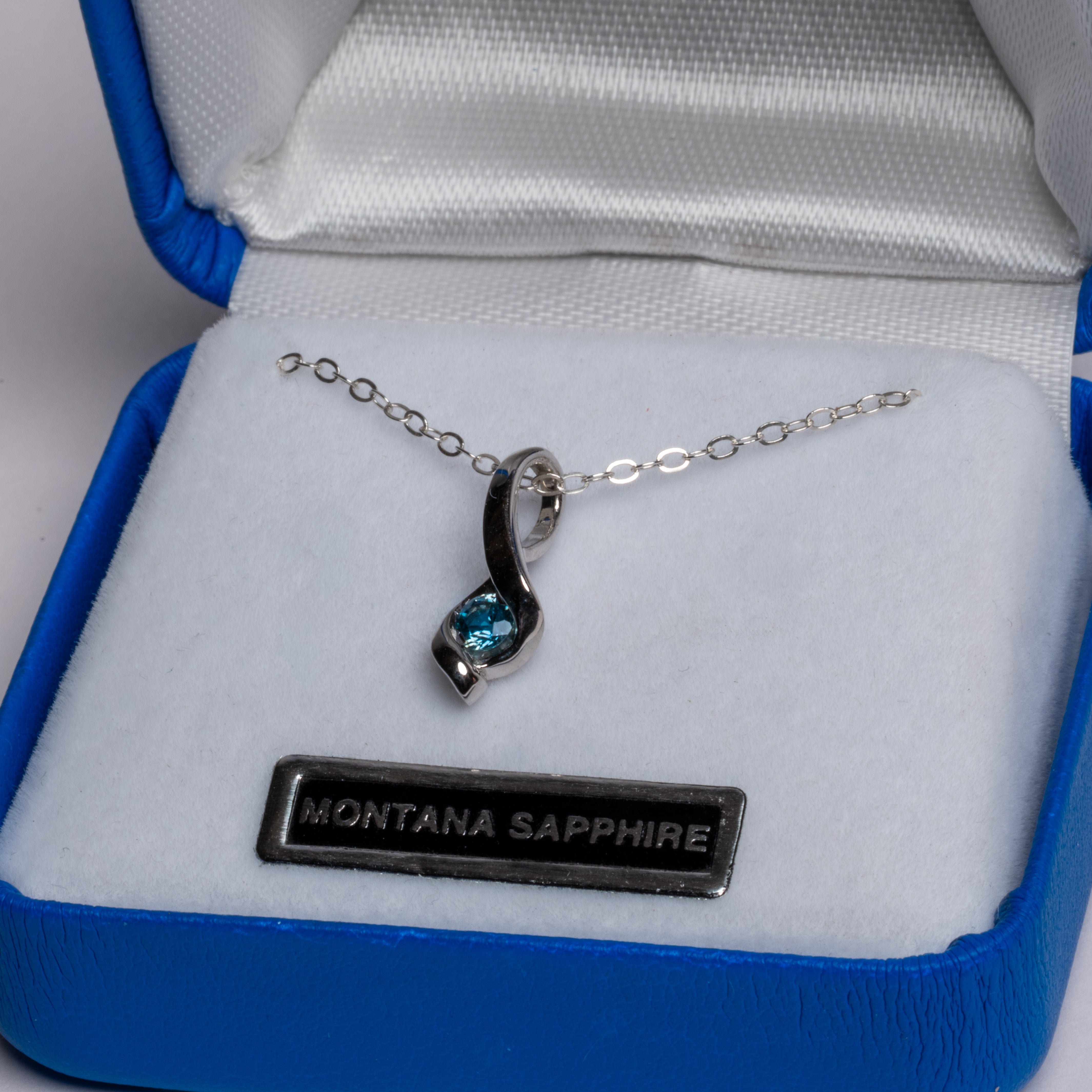 A silver-tone necklace with a twisted pendant featuring a round-cut blue Montana Sapphire gemstone, presented in an open blue jewelry box with a white interior. A silver nameplate in the box reads 'MONTANA SAPPHIRE'.