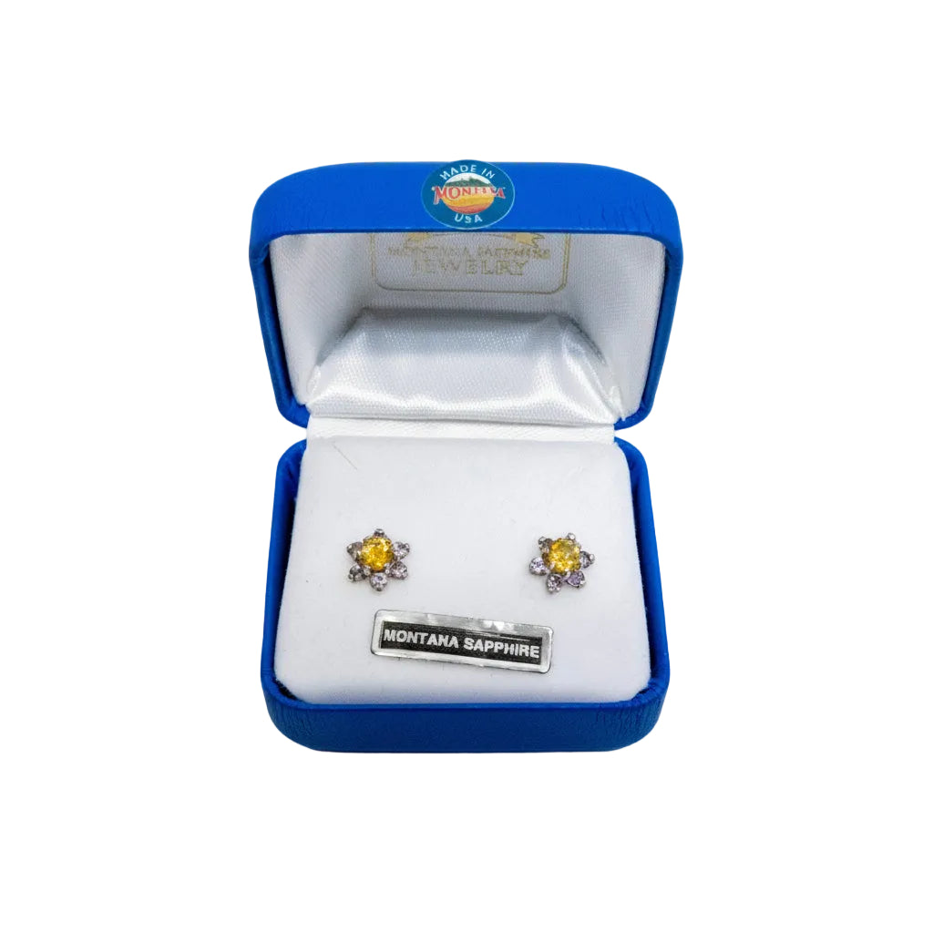 A pair of Montana Sapphire stud earrings with yellow center stones and silver outer stones, displayed in an open blue Montana Jewelry box on a white background.
