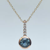 A rose gold necklace with a round-cut blue Montana sapphire gemstone pendant and a vertical pavé bar of small clear stones, shown on a light background.