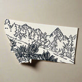 White dish cloth with black mountain and plant design on a light wooden surface.