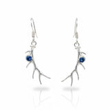 A pair of silver drop earrings shaped like antlers, each accented with a small round blue gemstone, shown on a clean white background with a soft reflection below.