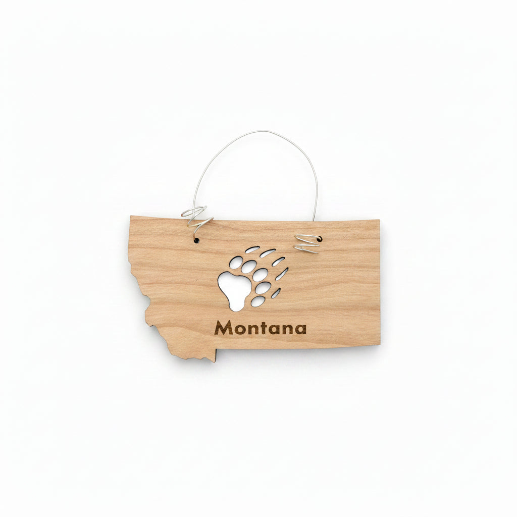 A small wooden ornament shaped like the state of Montana, hanging from a thin metal wire loop. The wood is light tan with a black bear paw print and the word "Montana" engraved on it.