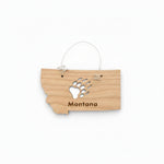 A small wooden ornament shaped like the state of Montana, hanging from a thin metal wire loop. The wood is light tan with a black bear paw print and the word "Montana" engraved on it.