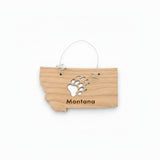 A small wooden ornament shaped like the state of Montana, hanging from a thin metal wire loop. The wood is light tan with a black bear paw print and the word "Montana" engraved on it.