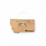 A small wooden Montana-shaped ornament or plaque with a black pinecone engraving and the word 'Montana' on a dark wood surface. A small white paper tag is attached at the top by a thin metal loop.
