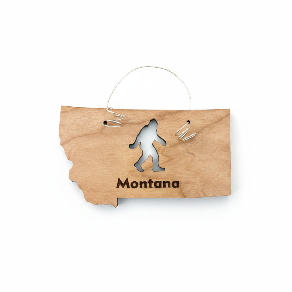 A wooden ornament shaped like the state of Montana, featuring a black engraved silhouette of Bigfoot and the word 'Montana' at the bottom. It is displayed on a dark brown wooden surface with a small white price tag attached at the top.