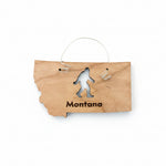 A wooden ornament shaped like the state of Montana, featuring a black engraved silhouette of Bigfoot and the word 'Montana' at the bottom. It is displayed on a dark brown wooden surface with a small white price tag attached at the top.