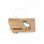 A small wooden ornament shaped like the state of Montana, engraved with the word Montana and a moose silhouette, hanging from a thin metal loop on a dark wood tabletop.