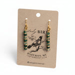 A pair of dangling earrings with green and gold spacer beads and gold hooks, displayed on a brown kraft paper card with 'Lady Bird' branding.