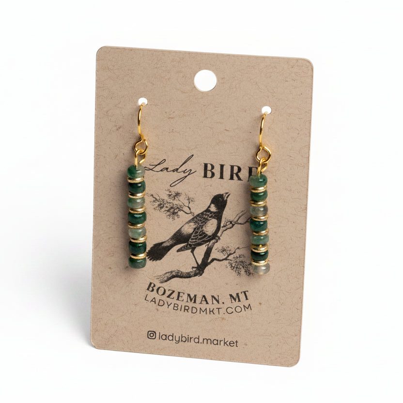 A pair of dangling earrings with green and gold spacer beads and gold hooks, displayed on a brown kraft paper card with 'Lady Bird' branding.
