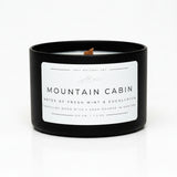 A black candle jar with a white label reading 'MOUNTAIN CABIN' and 'NOTES OF FRESH MINT & EUCALYPTUS,' sitting on a white background