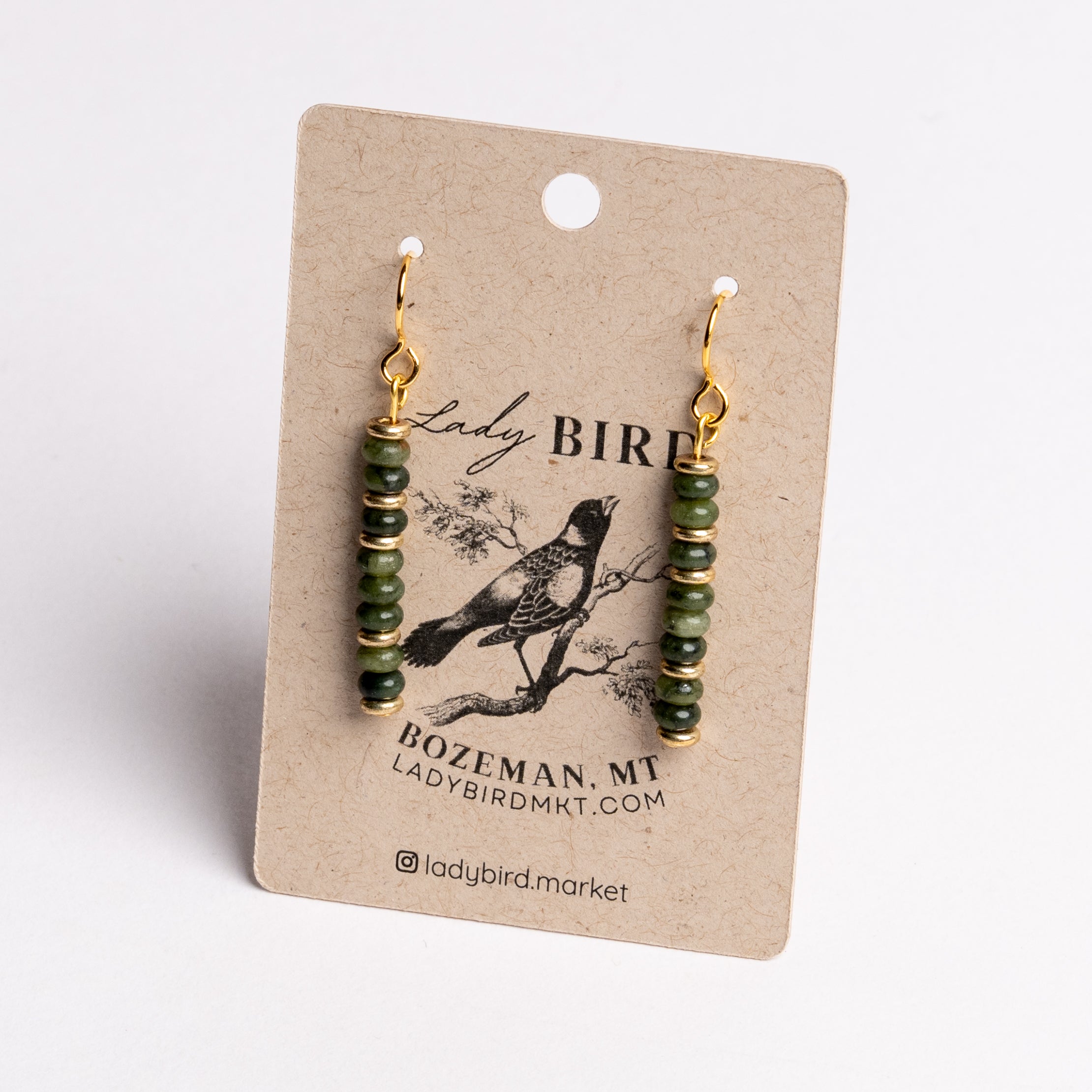 A pair of dangling earrings with green faceted beads and gold hooks, displayed on a kraft paper card printed with the brand name Lady Bird.