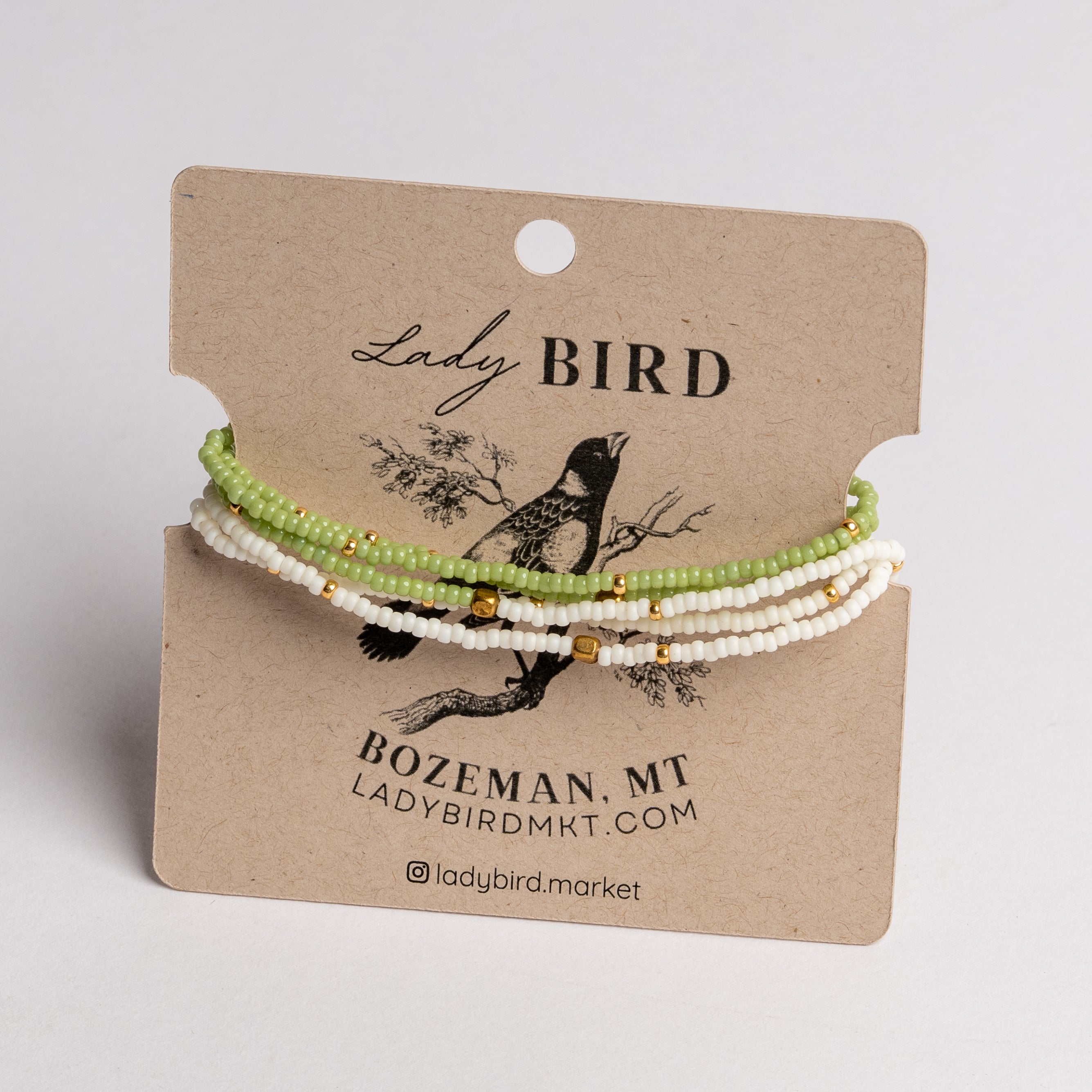 Three beaded bracelets in light green, white, and gold tones displayed on a brown kraft card labeled Lady Bird, photographed against a light neutral background.