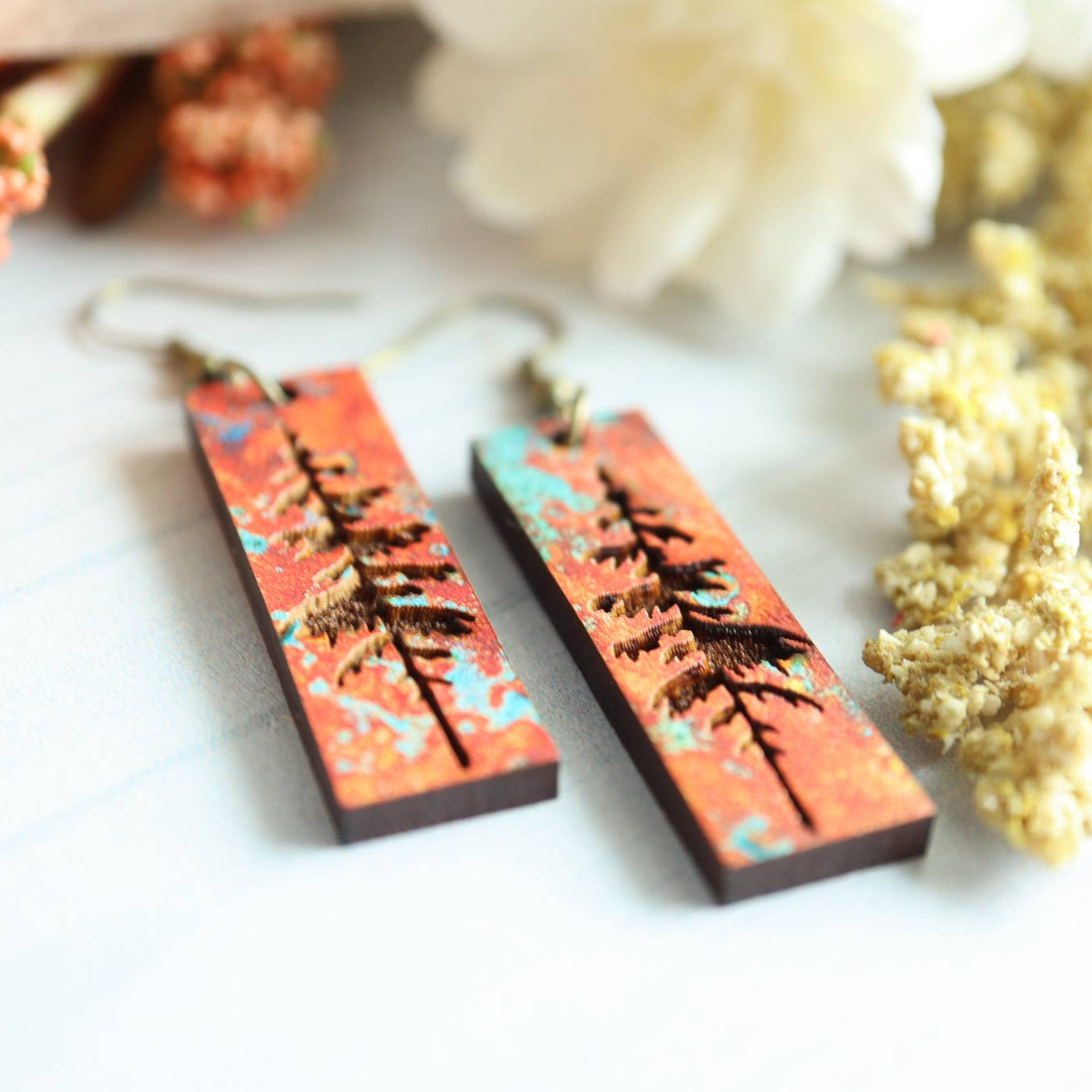Pair of earrings with tree design on a white surface with flowers in the background
