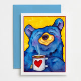 A greeting card featuring a colorful illustration of a blue bear holding a white mug with a red heart, placed in front of a blue envelope on a white surface.