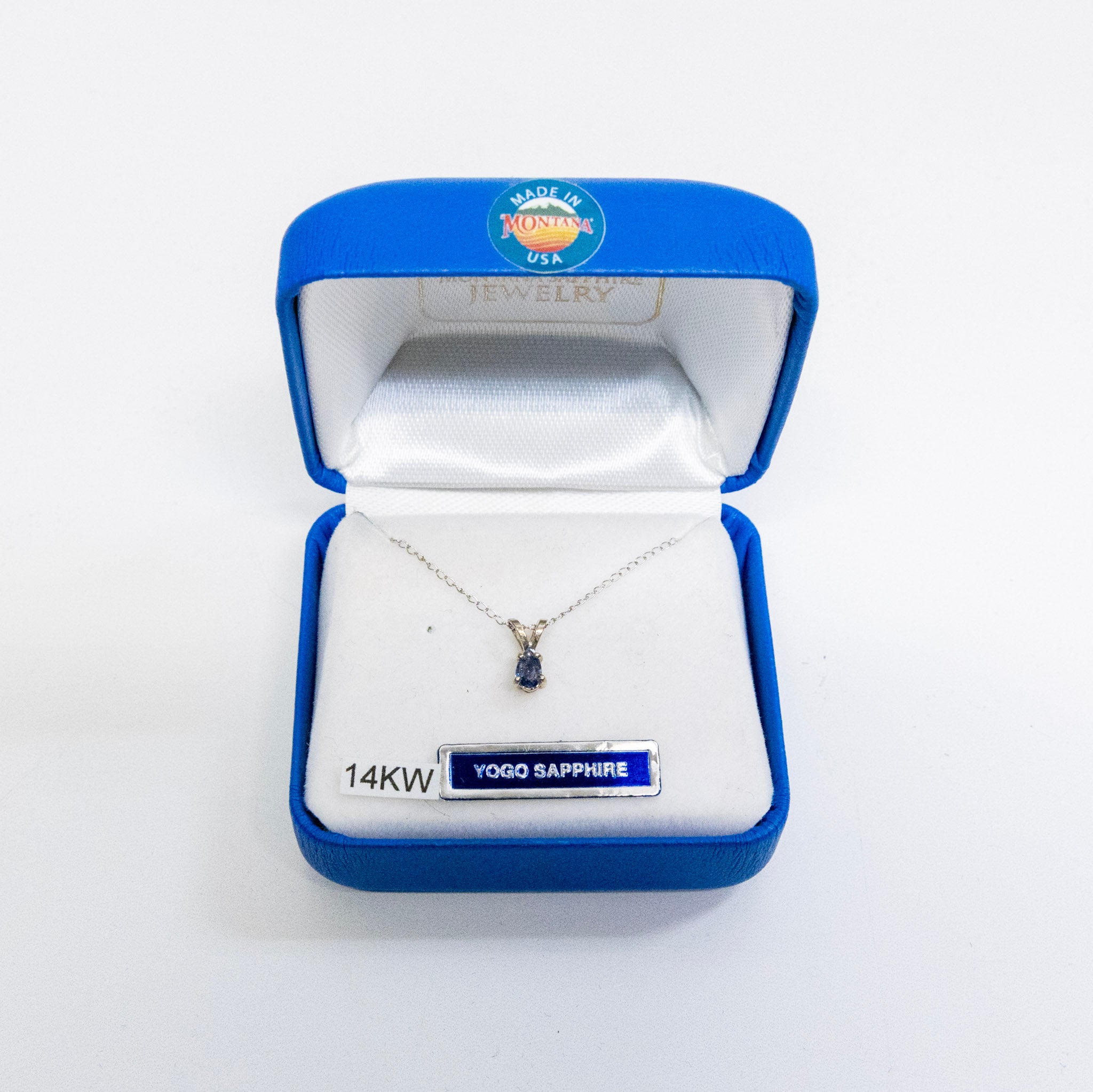 A silver necklace with a small blue sapphire pendant is displayed inside an open blue jewelry box with a white satin lining. The box has a 'YOGO JEWELRY USA' logo on the inside lid and a '14KW YOGQ SAPPHIRE' label on the front.