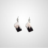 A pair of dangling earrings shaped like the state of Montana, featuring a printed landscape design with black pine trees against a pale pink and white sky, suspended from silver fishhook wires on a light gray background.