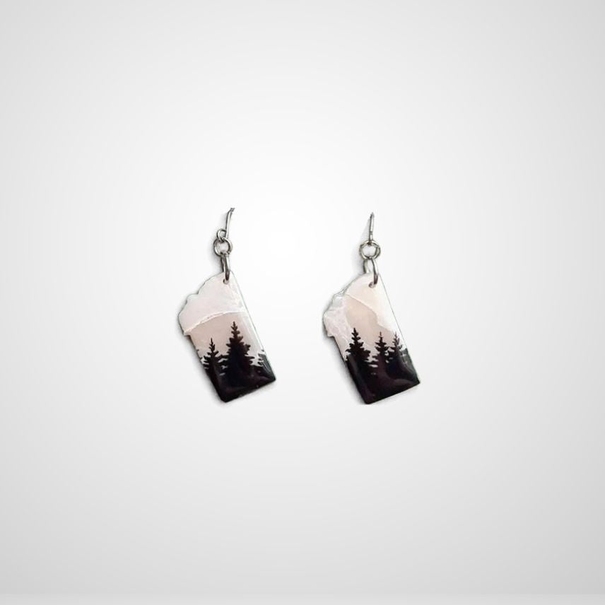 A pair of dangling earrings shaped like the state of Montana, featuring a printed landscape design with black pine trees against a pale pink and white sky, suspended from silver fishhook wires on a light gray background.