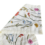 A folded cream-colored cotton muslin cloth with a colorful wildflower print, displayed on a white-washed wooden background.