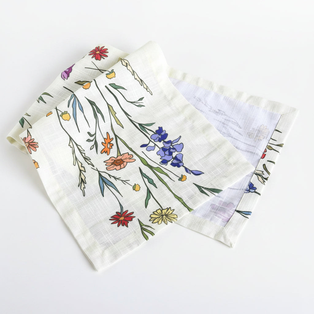 A folded white gauze bandana or napkin featuring a colorful hand-drawn style floral and botanical print, displayed on a white-washed wooden surface.