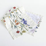 A folded white gauze bandana or napkin featuring a colorful hand-drawn style floral and botanical print, displayed on a white-washed wooden surface.