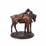 A bronze-finished sculpture depicting a cowboy riding a horse while another person stands beside it, mounted on an oval decorative base against a plain white background.