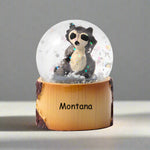 Snow globe with a raccoon figurine on a wooden base labeled 'Montana' against a white background