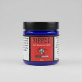 A jar of Natural Encouragement 'Ripped' sore muscles and joints cream, featuring a dark blue jar, black lid, and red label, set on a rustic wooden surface.