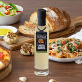 Bottle of Olivelle Roasted Garlic oil on a wooden table with food items like pizza, pasta, and bread.