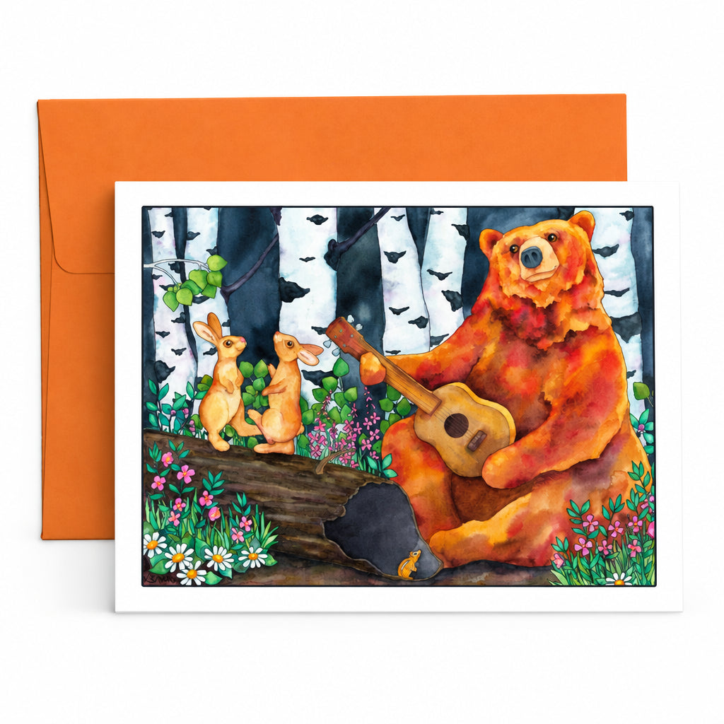 A square art print or greeting card featuring a painted forest scene with a bear playing guitar, two rabbits, birch trees, flowers, and a log, shown in front of an orange envelope on a white surface.