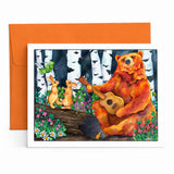 A square art print or greeting card featuring a painted forest scene with a bear playing guitar, two rabbits, birch trees, flowers, and a log, shown in front of an orange envelope on a white surface.