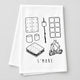 A white kitchen towel with black line art illustrations of s'mores (graham crackers, marshmallows, ice cream, s'more stack, campfire) and the text 'S'MORE' at the bottom, laid flat on a wooden surface.
