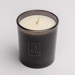 A single-wick scented candle in a dark smoky gray glass jar with a white wax fill and a light wood wick. The front of the jar features a frosted gray square logo containing the numbers '1889'.