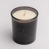 A single-wick scented candle in a dark smoky gray glass jar with a white wax fill and a light wood wick. The front of the jar features a frosted gray square logo containing the numbers '1889'.