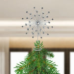 A silver metal Christmas tree topper with sparkling rhinestone-like beads, shown mounted on a green pine tree branch against a softly blurred indoor background.