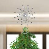 A silver metal Christmas tree topper with sparkling rhinestone-like beads, shown mounted on a green pine tree branch against a softly blurred indoor background.