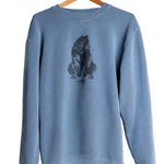 A dusty blue crewneck sweatshirt hanging on a wooden hanger against a white wall. The front of the sweatshirt features a dark gray watercolor-style graphic of a bear sitting on its hind legs, with small red text underneath