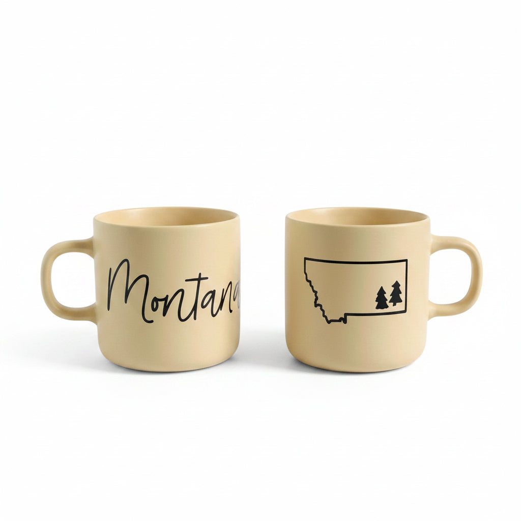 Two light beige ceramic mugs on a white wooden table, one printed with the word 'Montana' in black script and the other with a black outline map of Montana containing two pine trees.